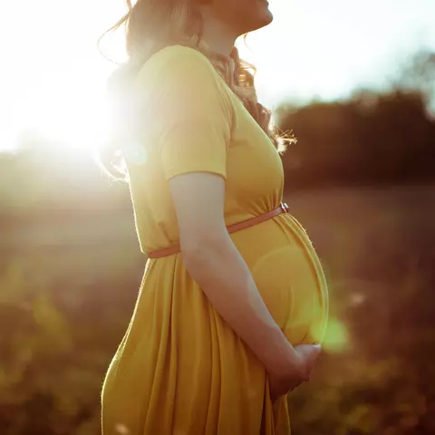 Schwangere Frau steht draußen ine einem Feld und hät ihren Bauch.